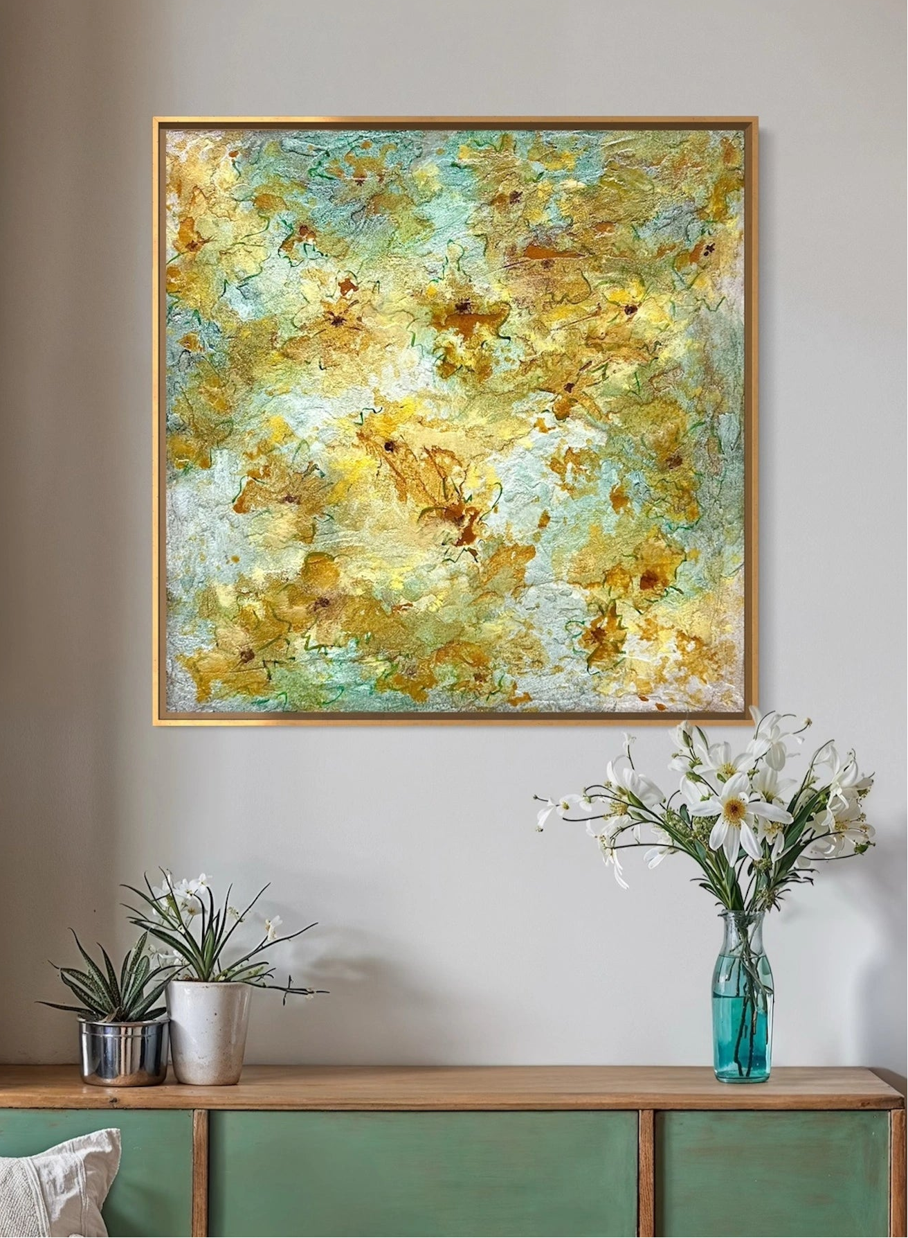 An abstract textured painting with gold, yellow, and mint green brushstrokes in a thin gold frame, displayed on a light gray wall above a wooden console table with green drawers, white flowers, and small potted plants.